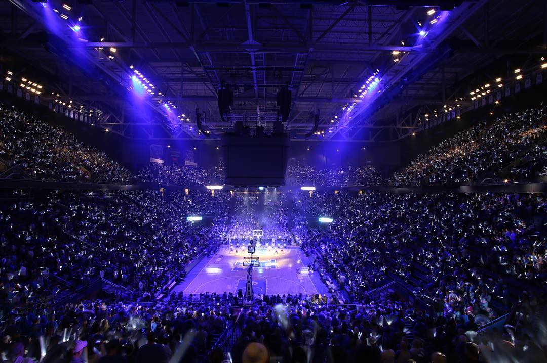 A packed indoor arena with blue lights illuminating a basketball game.