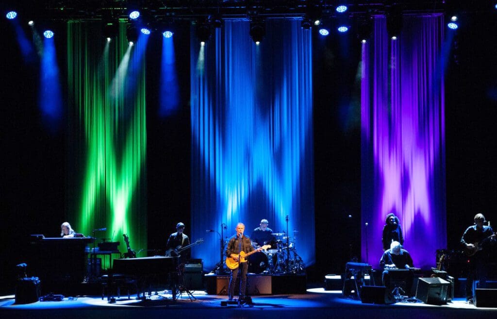 A band performs on stage with colorful blue, green, and purple lighting.