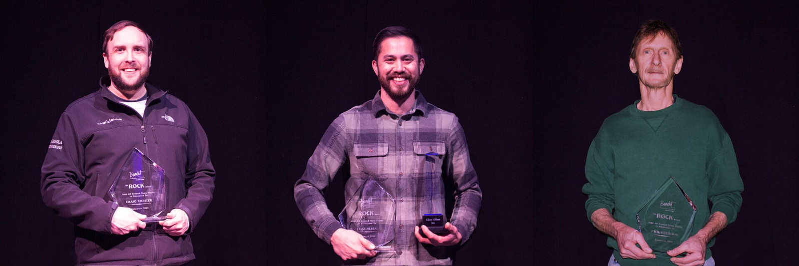Smiling man in a plaid shirt holding a microphone on stage.