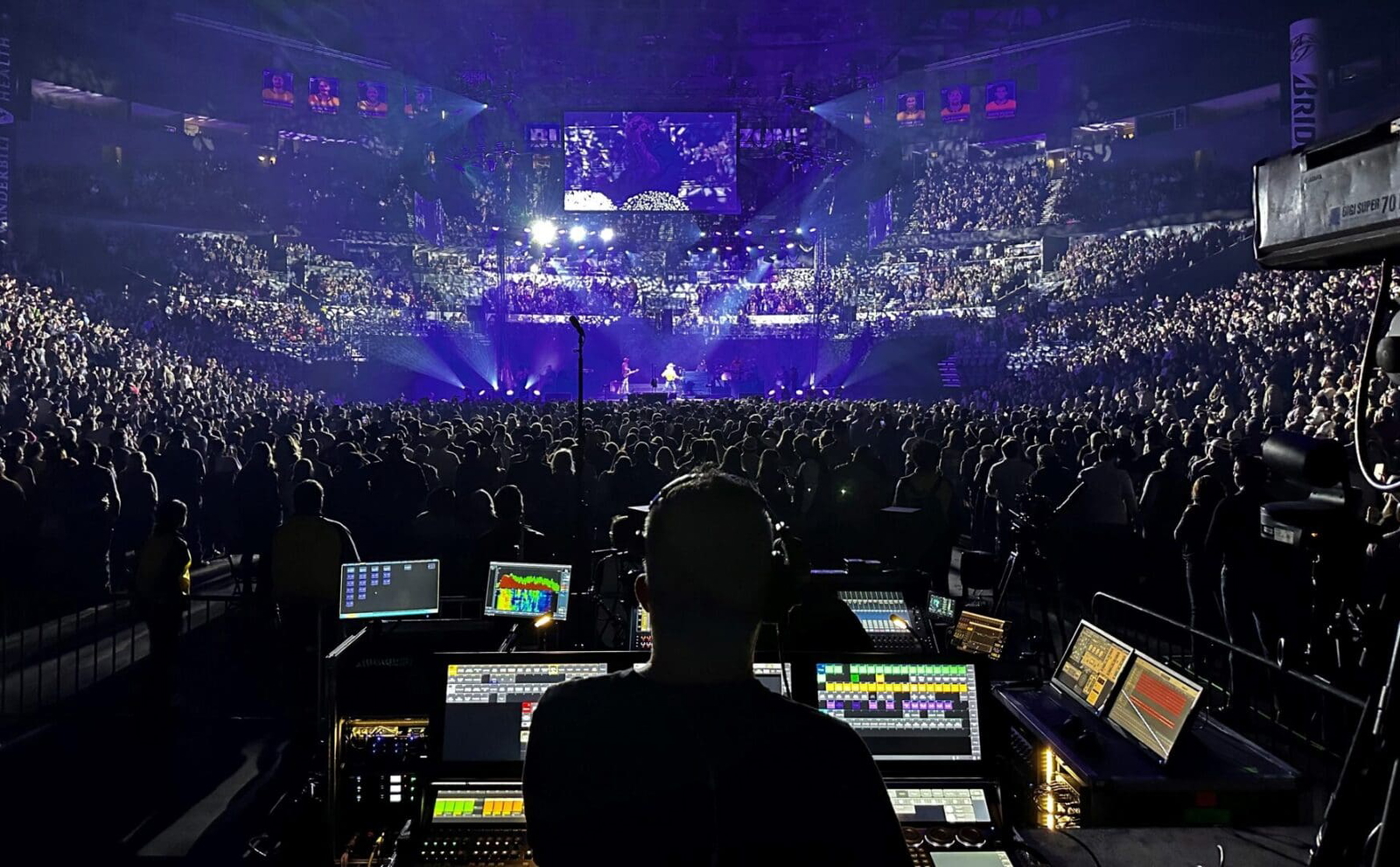 A sound engineer manages audio at a packed indoor concert with vibrant purple lighting.
