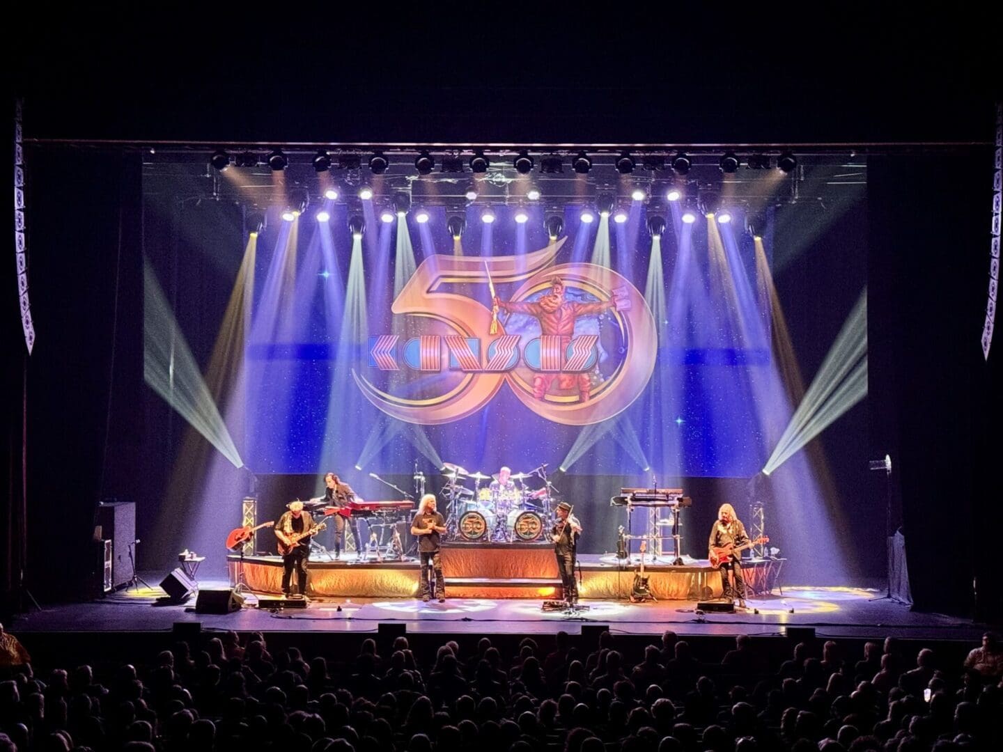 A live band performs on stage with vibrant lighting and a large logo backdrop.
