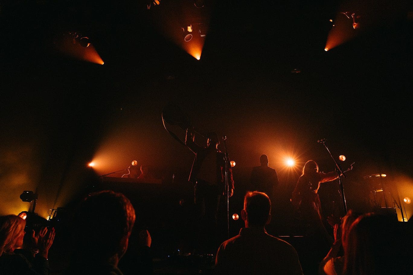 Silhouetted band performing under warm stage lights in a dark venue.