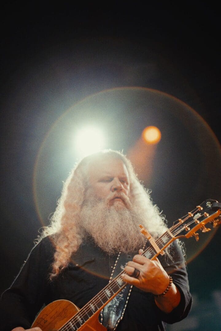 A bearded man playing guitar on stage with bright lights behind him.