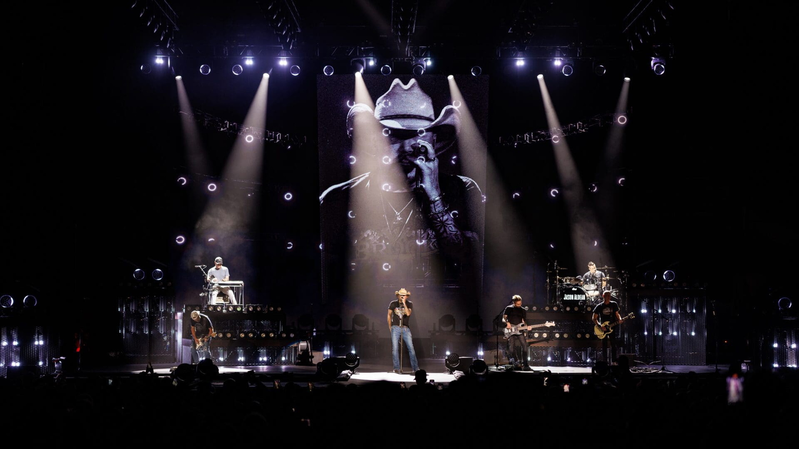 A dramatic live concert with a central singer illuminated by spotlights and a large black-and-white face projection.