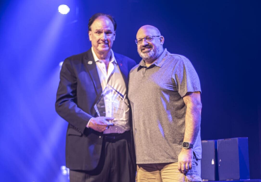 Two men standing on stage with award and microphone, smiling. Two men standing on stage with award and microphone, smiling.
