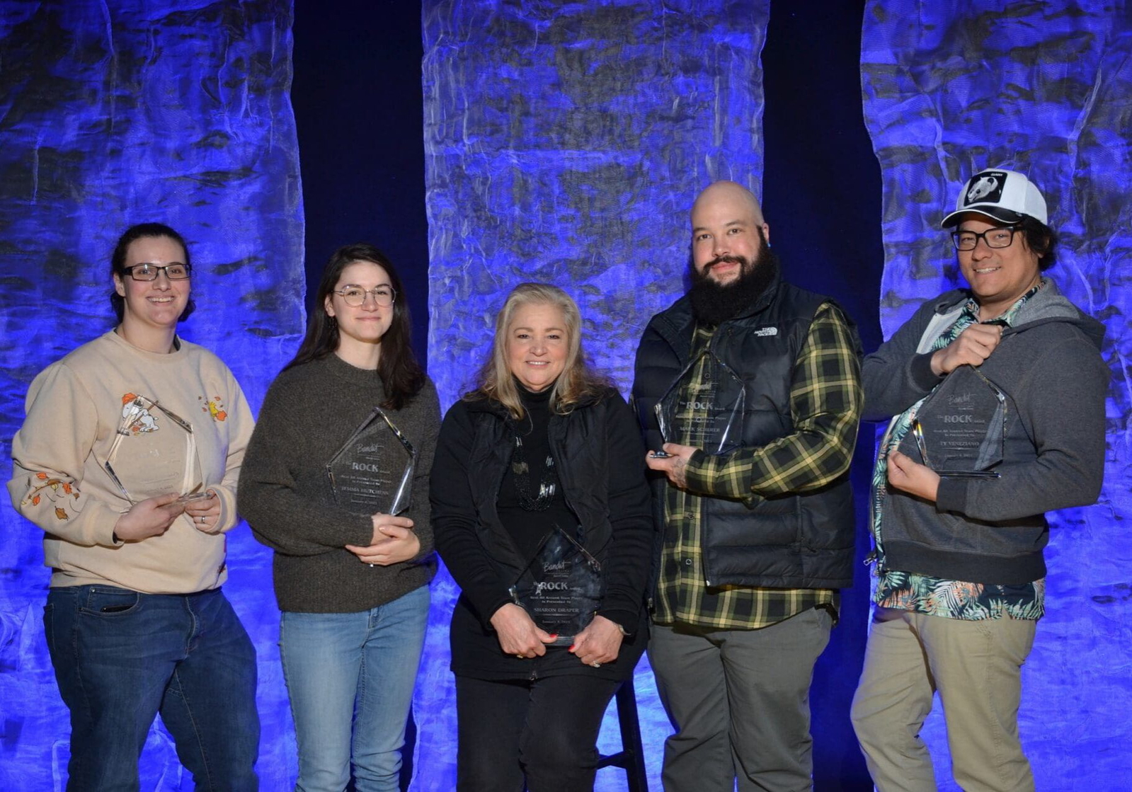 Five people posing together against a stone wall with blue lighting. Five people posing together against a stone wall with blue lighting.