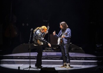 Two musicians perform on stage with guitars under dark lighting.