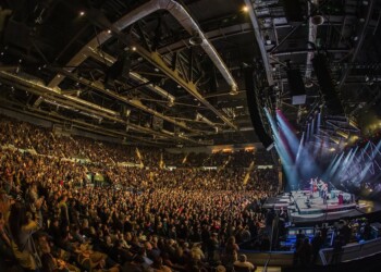 Massive crowd enjoying a concert in a large indoor arena with bright stage lights.