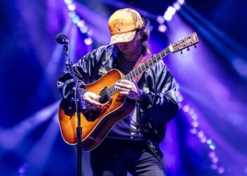 Musician passionately playing acoustic guitar on stage with vibrant purple lighting.
