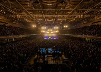 A packed indoor arena with bright stage lights and a large crowd.