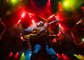 Photo: Jesse Faatz Guitarist passionately playing on stage with vibrant lights.