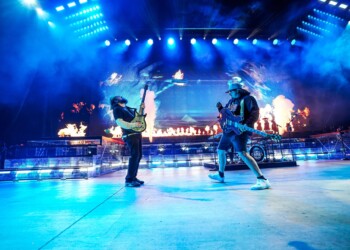 Two guitarists perform energetically on a brightly lit stage with a large audience.
