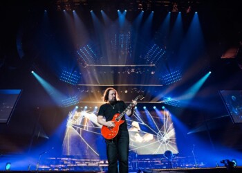 Guitarist performing on a large, illuminated stage with a massive screen behind.