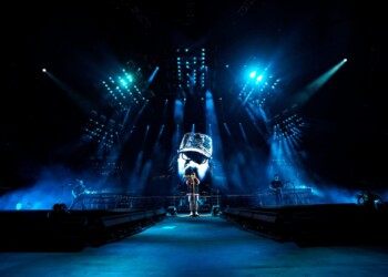 A performer stands on stage under blue lights with a futuristic backdrop.