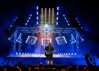 Musician performs on stage with bright blue lights and audience in front.