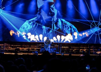 A performer in a blue, illuminated costume dances on stage with a large, glowing backdrop.