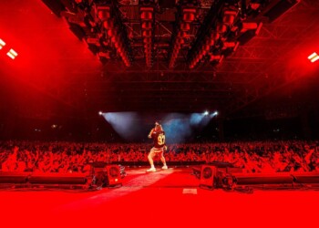 A performer on stage under red lights with a large audience.