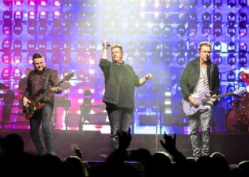 Band performing energetically on a brightly lit stage with a cheering crowd.