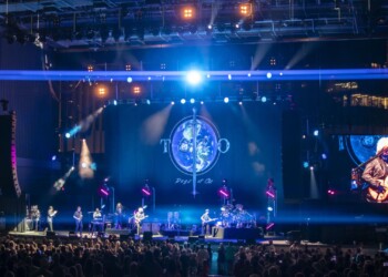 A band performs on stage with a glowing clock backdrop and vibrant lights.