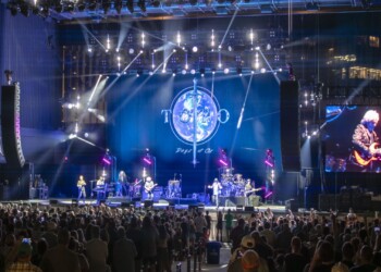 A band performs on a large stage with vibrant lighting and a crowd watching.
