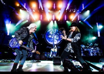 Two guitarists perform energetically on stage under vibrant lights.