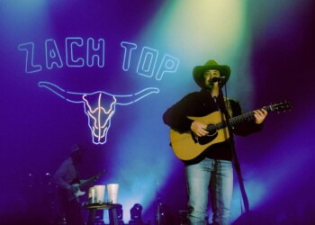 Musician playing guitar on stage with neon 'Beach Top' sign and colorful lights.