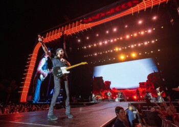 Musician performing energetically on stage with guitar under bright lights.