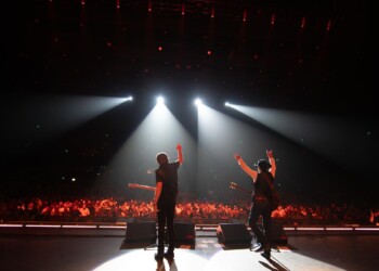Performers on stage illuminated by spotlights with audience in the background.